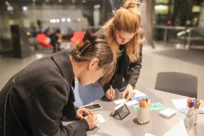 Two young women writing on a table in the atrium of the MIT List Visual Arts Center