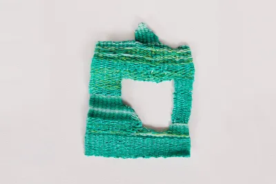 The fibers of this small square shaped weaving are shot through with variegated green threads in shades ranging from mint to lime to teal. The center has a hole in the shape of a square missing its lower left corner through which the white wall is visible.