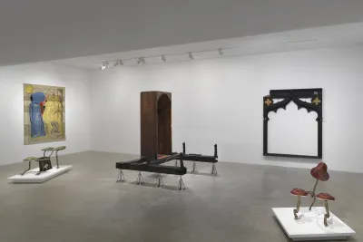 A white gallery with five artworks on display. On the left wall hangs a gold tapestry depicting two figures, one standing and one kneeling. Two small creature-like sculptures made of metal and fabric sit on low pedestals. In the center of the room is a vertical wooden bathtub sculpture on metal supports. A large black frame-like wooden sculpture hangs on the back wall.