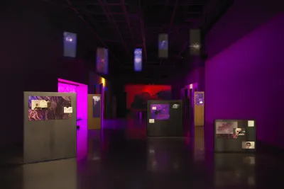 A dark room with purple walls is occupied by video projections and illuminated artworks on the floor and ceiling, creating an effect like overlapping windows on a computer screen. A purple glow comes from a tinted window on the left side. Freestanding panels are scattered throughout the room, each with colorful abstractions and other images mounted to them. Blue banners with abstracted plant designs hang from the ceiling and a red video projection, showing a person lying on their back, glows on the far wall