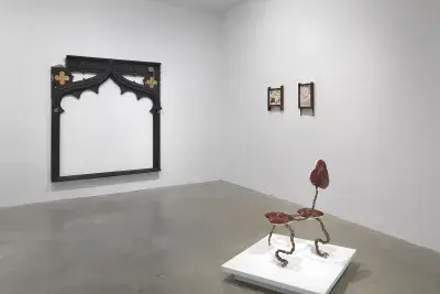 A white gallery with four artworks on display. On the left a large black frame-like wooden sculpture hangs on the back wall. To the right of the frame-like sculpture, two small drawings hang in wooden frames. In the foreground, a small creature-like sculpture made of metal and fabric sits on a low pedestal.