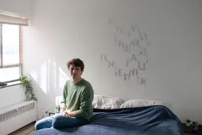 A man wearing a green sweater is sitting on a bed with navy sheets in front of a stenciled artwork on the white wall behind. The bed is to the right of a window with a plant on the windowsill.