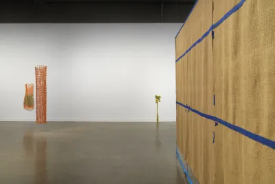 On the left, two cylindrical orange net-like sculptures are displayed. One is much smaller than the other; the smaller one incorporates an inner green component. On the far right, a large brown tapestry hangs from the ceiling. Blue fiber strips grid the piece with some of the strips dangling onto the floor. In the background on the right, a small, yellow caution tape fiber sculpture is bunched up dangling off the wall and onto the floor.