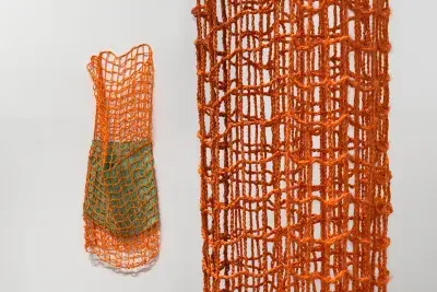 A close-up view of a woven netlike structure of a hanging textile artwork. The artwork forms a kind of dense cylinder, with several overlapping layers of material. On the left of the image, another orange netlike artwork hangs on the wall encasing a smaller green fiber cylinder inside of it.