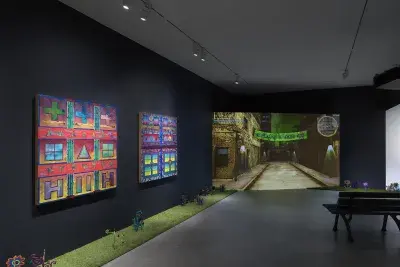 A dark, black-walled gallery space with artworks on display. On the left wall, two‬ ‭ paintings hang in a row depicting bright, geometric forms. In the back of the gallery, a‬ ‭ large screen illuminates the space displaying digitally rendered footage of a dark city‬ ‭ street with a green banner hung between buildings. A park bench faces the screen. A‬ ‭ strip of astroturf lines the floor around the gallery, with painted wooden flowers emerging‬ ‭from it.‬ ‭