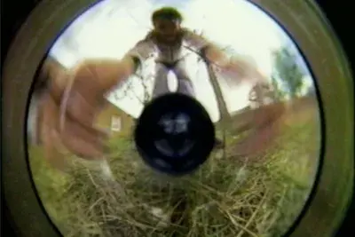 A round, distorted image of a woman set within a rectangular frame. A smaller black circle occupies the center of the frame, like an eye or lens around which the image revolves. The woman stands and reaches down toward the camera; the perspective makes it seem like her hands are as big as the entire rest of her body . The texture of grass is visible in the bottom portion of the image, while a building, trees, and sky are visible in the background.