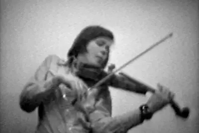 A woman in a button-up shirt plays a violin. She leans slightly backwards or to the side, as if caught in the middle of full-body swaying or dancing. The image is grayscale and distinctly low resolution.