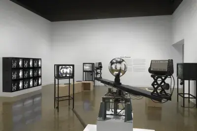 In the foreground, a sculptural apparatus comprises two cameras on the ends‬ ‭ of a single rotating arm, each pointed inwards at a central mirrored sphere. A feed from‬ ‭ the cameras are routed to a nearby monitor. In the background, other video monitors‬ ‭ are visible, including a grid of nine showing the same abstract black-and-white pattern,‬ ‭along with exhibition wall text.‬ ‭