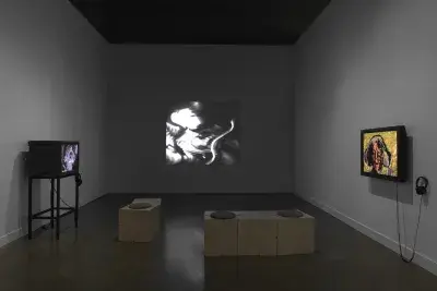 Three videos are shown in a gallery space. At center, a distorted image of a‬ ‭ person playing the violin is projected on the wall: the bow is curved and the person‬ ‭ indistinct, hinting at its electronic manipulation. At right, a flat monitor shows an image of‬ ‭ an older woman against a yellow background; portions of the background show through‬ ‭the face. At right, a boxy video monitor sits on a stand.‬ ‭