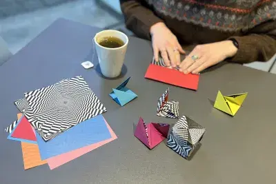 Two hands folding colorful geometric paper. There is a white mug of tea to the left side of the hands and a stack of unfolded paper in the foreground.