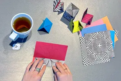 Overhead view of two hands folding colorful geometric paper. There is a white mug of tea to the left side and a stack of unfolded paper to the right.