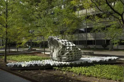 A large rock sculpture rests on a long strip of stone cylinders in a‬ ‭ greenspace. The rock has a series of holes removed from its core, with the‬ ‭ cylinders that were removed making up the strip it rests on. Trees, a pathway,‬ ‭ and a gray building reside in the background.‬