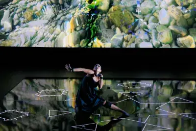 Video still of a performance showcasing a woman spotlighted on stage with her head tilted up dancing. There is a bright iridescent pattern projected on the wall behind.