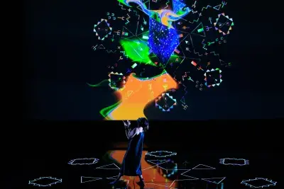 Video still of a performance showcasing a woman spotlighted on stage with her head tilted up, arms raised and palms facing up. A brightly colored geometric pattern in the background is juxtaposed by a black wall.
