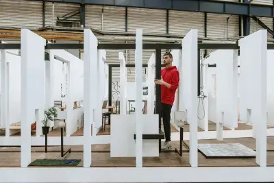 A series of fragmented white walls divides a living space with wooden floors, furniture, and house plants throughout. Each section of wall is connected by black beams. A man in a red sweatshirt stands in the center of the frame, grasping one of the black beams.