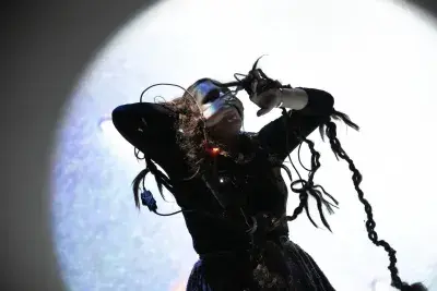 Loose braids of dark hair dangle from the hand and head of a performer‬ ‭ wearing a shiny silver mask. A loop of wire curls around her other arm and behind her‬ ‭ back. The boundary between the hair, the electronics, and her body is blurry and‬ ‭ shrouded in shadow. In the background is a bright projection of a pixelated blue circle.‬ ‭