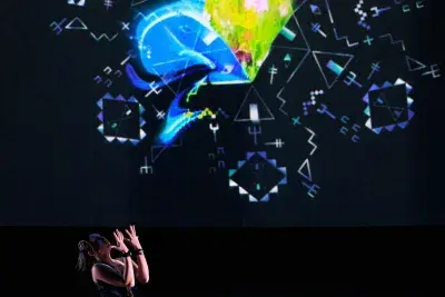 Video still of a performance showcasing a woman spotlighted on stage with her head tilted up, arms raised and palms facing up. A brightly colored geometric pattern in the background is juxtaposed by a black wall.