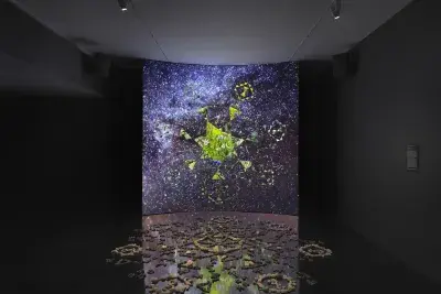 A dark gallery space features a large screen displaying a six-sided star at its‬ ‭ center. Within the star, video footage of green stones is projected. Surrounding the‬ ‭ star, six inward-facing triangles form a pattern, while geometric shapes swirl in the‬ ‭ background alongside footage of stars and the night sky. In front of the screen, a‬ ‭formation of stones rests on the reflective black floor.‬ ‭