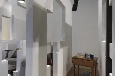 A series of fragmented white walls divides a living space with wooden floors, furniture, and house plants throughout. A wooden nightstand with a stereo and a stack of books is at the center of the frame. Each section of wall is connected by black beams.