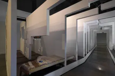 A walkway surrounded on both sides by a series of white walls that segments what appears to be a living space. A charging smartphone is visible on top of a bed in the bottom lefthand corner.