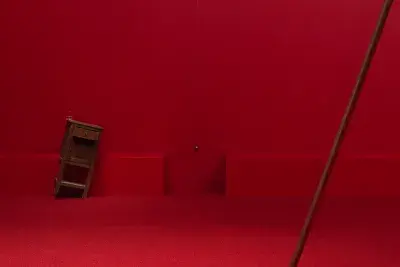 A wooden cabinet standing on a angle tilted to the right in a completely red painted room with red carpeting, next to a metal sphere hanging on a string suspended from the ceiling. Part of a copper poll is angled to the right in the right hand side of the frame.