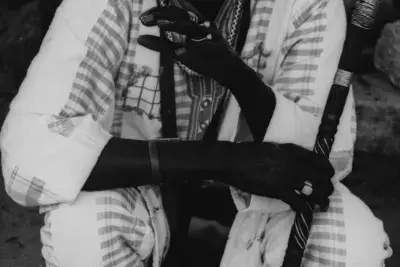 Black and white video still of a man sitting down holding a scepter wearing matching patterned clothing.