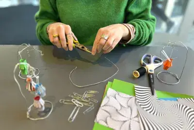 Two hands bending wire with wire cutters. There is a small mobile with a green paper lantern to the left side of the hands, a group of paper clips and a stack of unfolded paper in front of the hands, and scissors with another wire mobile to the right of the hands.