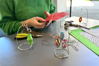 Two hands folding a red piece of paper. There is a small mobile with a green paper lantern to the left side of the hands, a group of paper clips and a stack of unfolded paper in front of the hands, and scissors with another wire mobile to the right of the hands.