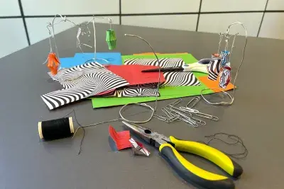 There are three small wire mobile's with a green, orange and multicolor paper lanterns on a gray table. There is a group of paper clips, a stack of unfolded paper, and a pair of bright yellow wire cutters in the foreground.