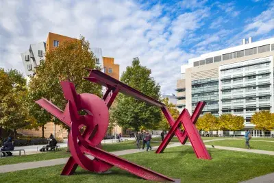 Angled view of a bright red steel sculpture on a green lawn on a sunny day with blue sky and puffy white clouds.