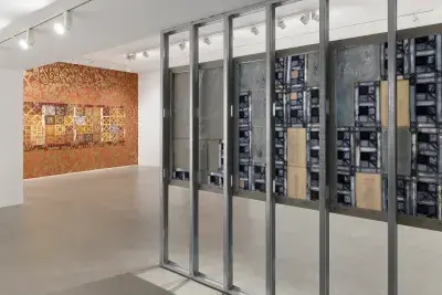 A gallery entryway with scaffolding on the right and the back of a blue patchwork-style painting in view. In the background, a painting hangs on an orange wall adorned with gold spray-painted motifs.