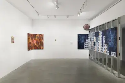 A blue cinder block-patterned painting wraps from scaffolding to the wall; a painted mirror and three other paintings hang throughout the gallery.