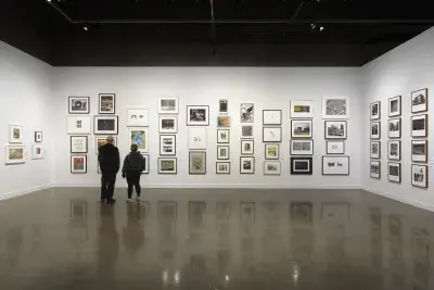 Installation view with framed artworks hanging on the walls salon style and two people standing beside each other in the center.