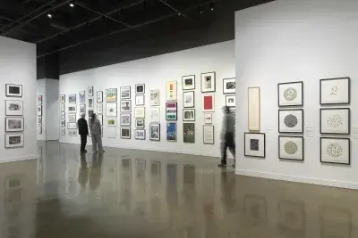 Installation view with framed artworks hanging on the walls salon style and three people standing looking at the art on the walls throughout the room.