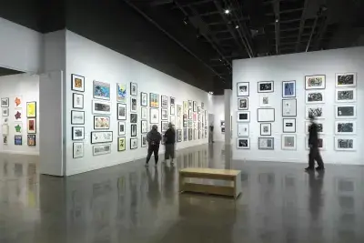Installation view with framed artworks hanging on the walls salon style and four people standing looking at the art on the walls throughout the room.