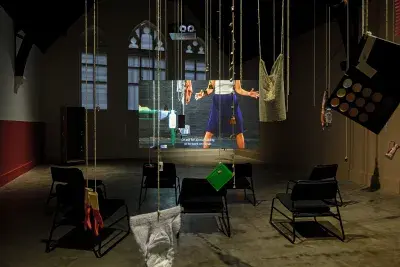 Dark open space with a video projected on a large standing screen in the back of the space. There are a collection of everyday items hanging from strings throughout the space.