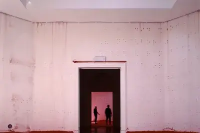 An open white-walled room with braille raised bumps across the walls. The silhouettes of two people can be seen through the dark doorway in the center of the image.