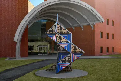 Large brightly colored geometric sculpture in the middle of a grass lawn in front of a brick building with a concrete dome awning.