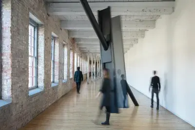Long rectangular space with light wood floors and a series of windows on the brick wall at the left side of the frame. There are large dark metal beams suspended in the middle of the space with people walking around the sculpture.