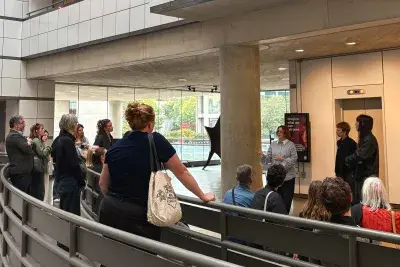 Crowd of people standing in a circle in an atrium listening to one person speak.