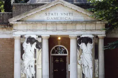 A classical stone building featuring tall columns supporting a grand entrance. There are two human figure statues on either side of the doorway. The text on the building facade reads, &quot;STATI VNITI D'AMERICA&quot;
