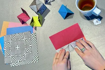 Overhead view of two hands folding colorful geometric paper. There is a white mug of tea to the left side and a stack of unfolded paper to the right.