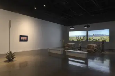 Inside a dimly lit gallery space, two projections sit behind a rocket test installation containing sandbags and canisters. In the foreground to the left is a metal sign inside an agave plant and a blue frame with pink paper mounted.