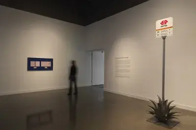 In a white gallery space, a blurred figure walks by a blue frame hung on the wall with 5 pink papers mounted inside. To the right is a Pasadena bus stop sign sitting amidst a metal agave plant.