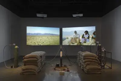 An installation of a rocket test containing sandbags, wiring, and canisters rest in a gallery space in front of two projections depicting people sitting in a desert landscape.