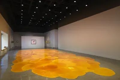 Gallery with an expansive, vibrant orange pool of melted pine rosin that spreads across the floor. Framed artworks hang on the back walls, while a window allows for warm sunlight to flood into the space.