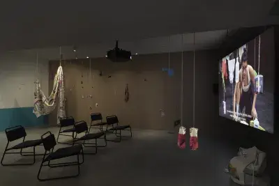 Dark open gallery with a video projected on a large standing screen on the right side of the frame and six black chairs in two rows in the center of the room. A tan canvas bag sits on the floor to the right of the screen. There is a collection of everyday items hanging from strings throughout the space. Two bright red rubber gloves hang in the foreground.