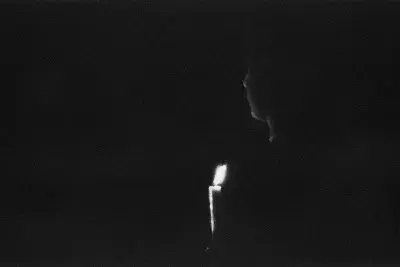 A black-and-white photo of a figure holding a candle in complete darkness. The image is predominantly black, with a lit candle at the bottom center of the frame. A faint silhouette of the figure is partially illuminated by the candlelight.