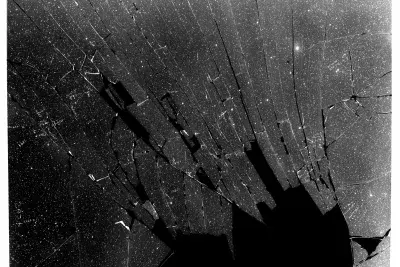 Black and white photograph of a shattered glass plate with a large black hole in the bottom right corner.