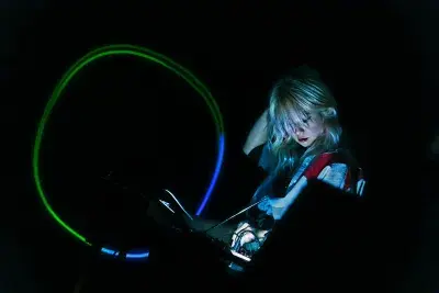 Illuminated female figure with platinum blonde hair in front of a DJ table, with a blue and green light circle to the left of the frame in front of a black background.